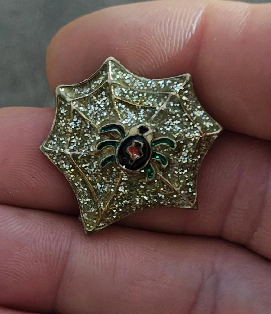 Gold spider web enamel pin with glitter details, black spider and red oval center, green accents
