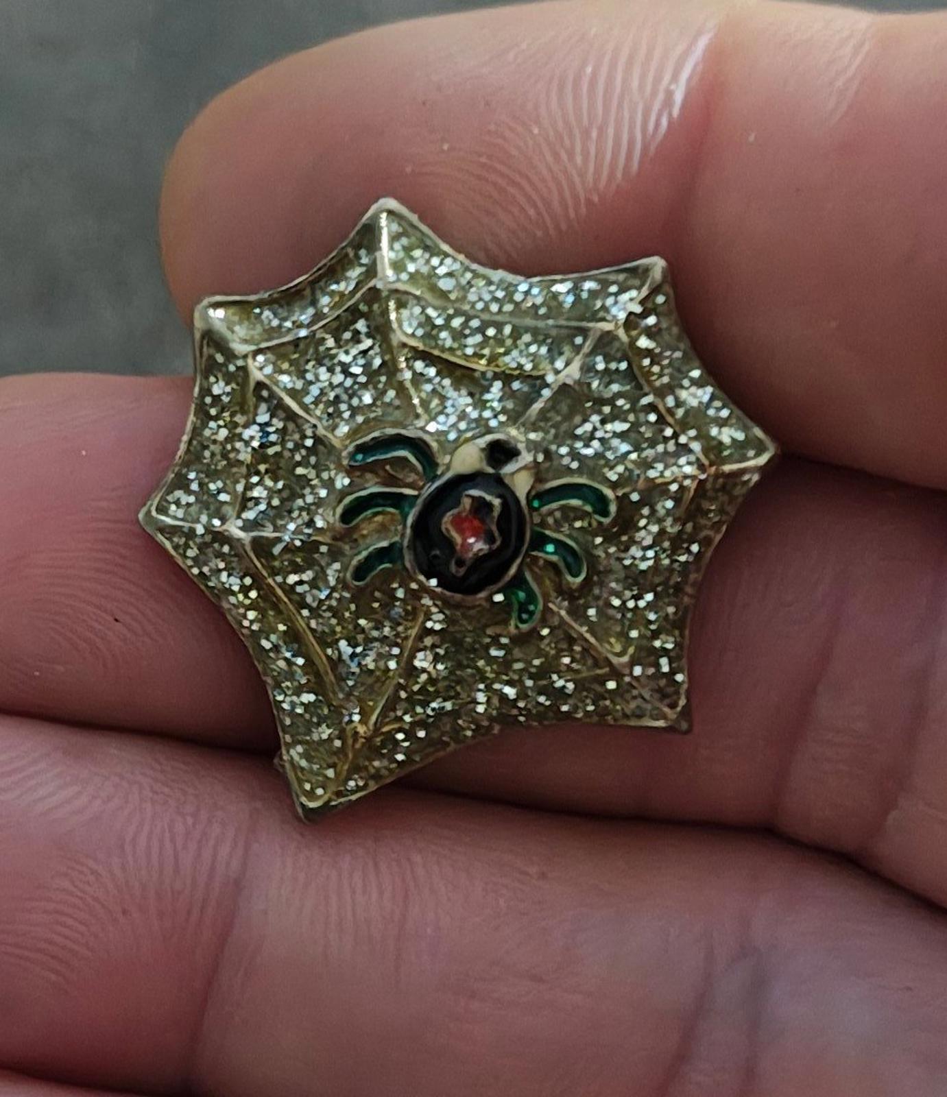 Gold spider web enamel pin with glitter details, black spider and red oval center, green accents