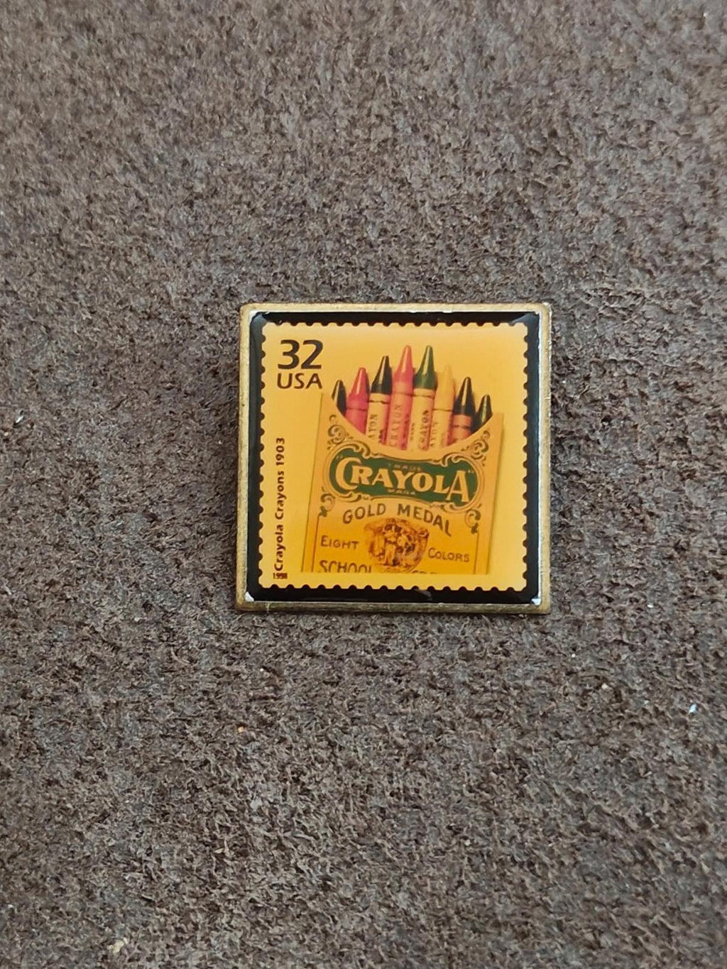 Square gold-tone Crayola stamp pin on yellow background with colorful crayons and “Gold Medal”