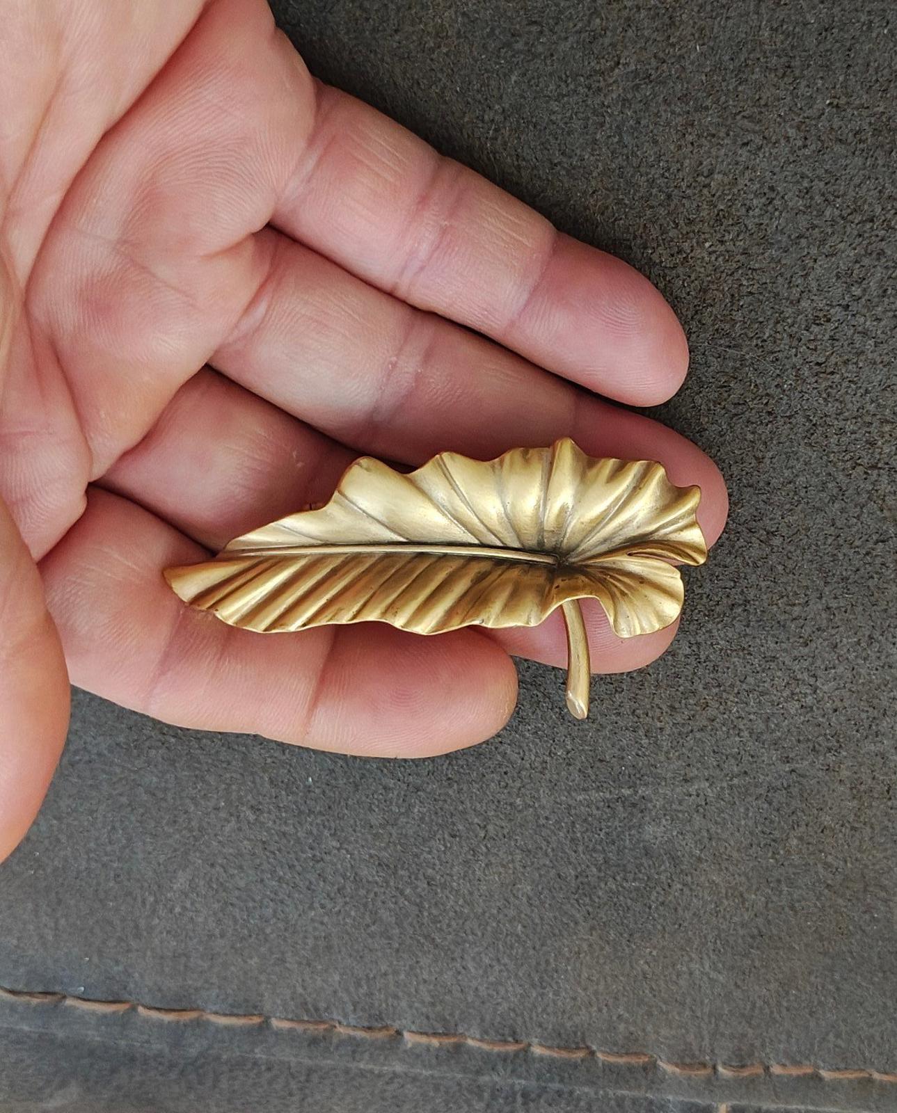 Gold-tone leaf brooch pin with textured ridges and stick pin, held in hand.