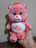 Pink plush bear with smiling face and heart belly emblem, held in a hand