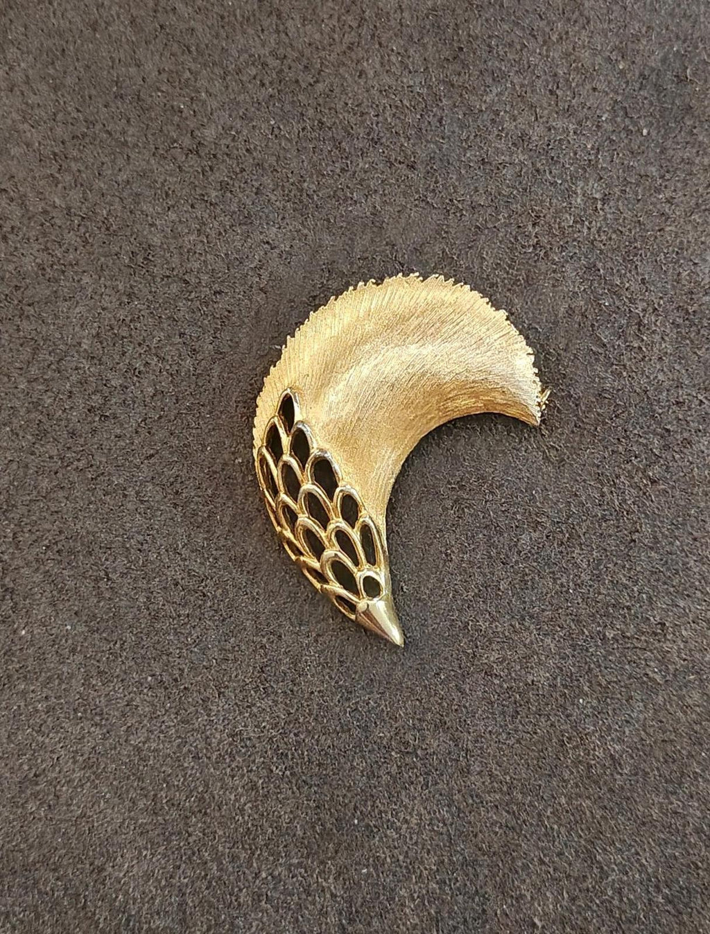 Gold tone feather plume brooch on dark gray background, curved top and pointed tip