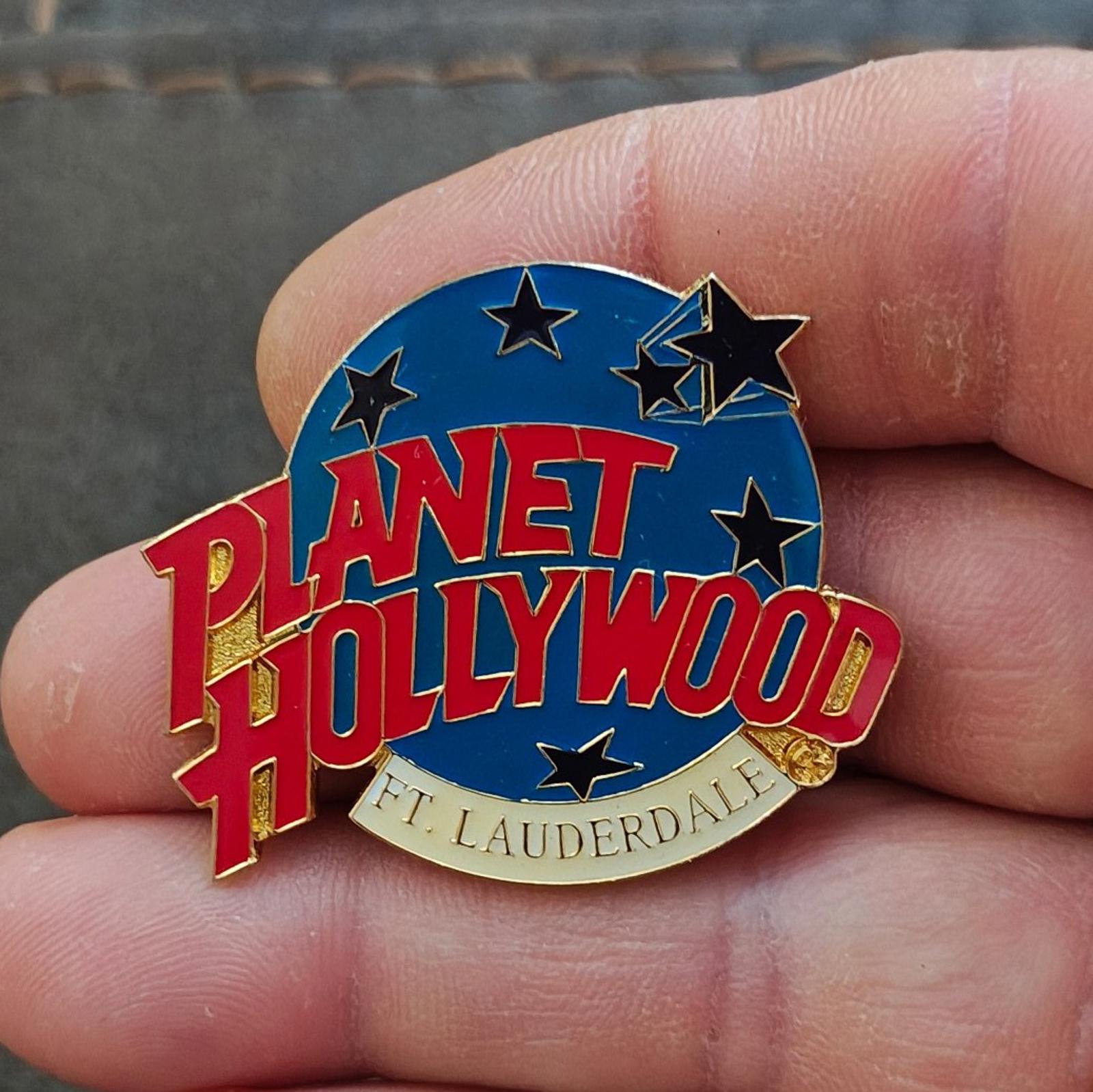 Blue enamel lapel pin in hand, red Planet Hollywood lettering, star accents, gold edge.
