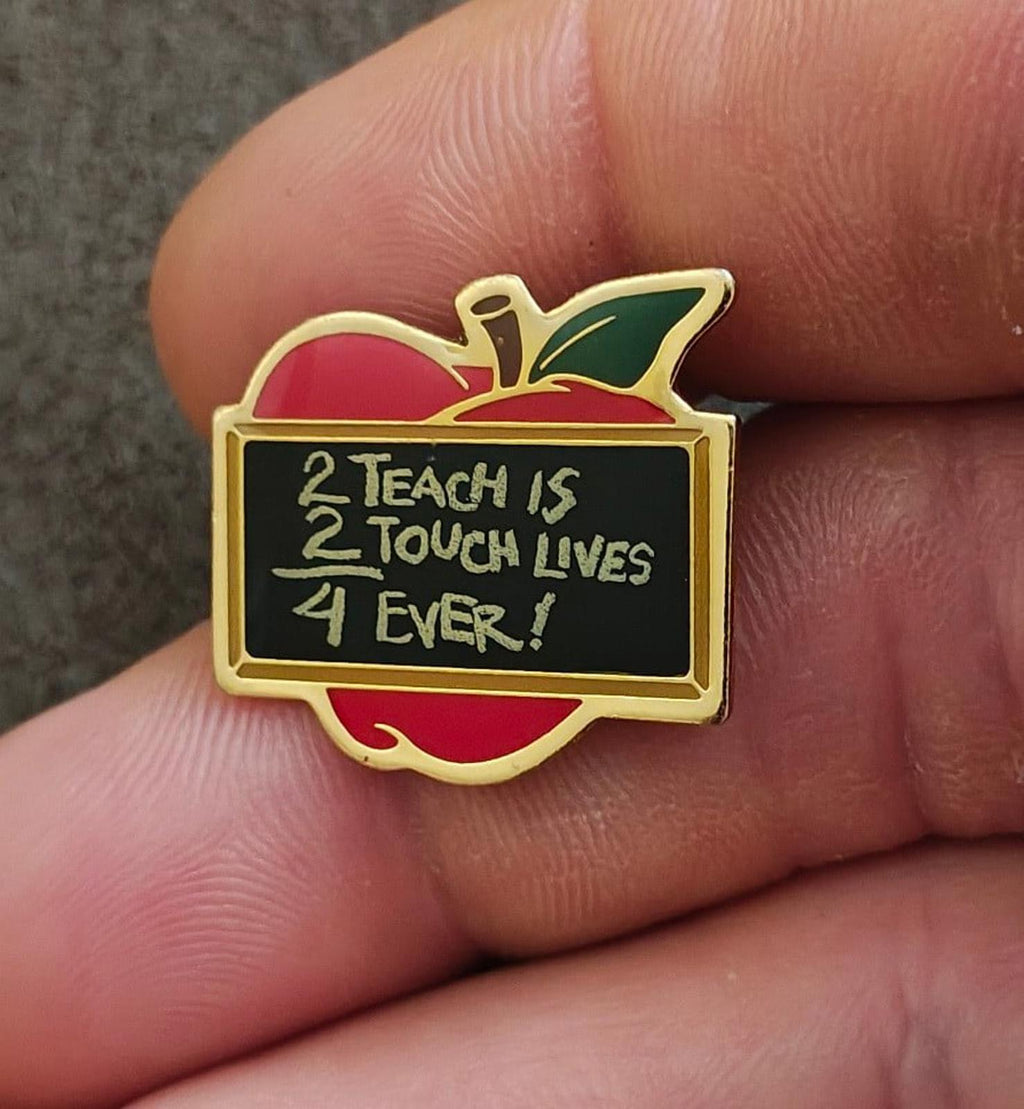 Gold-tone apple teacher pin with red and green enamel and black panel reading “2 Teach is…”