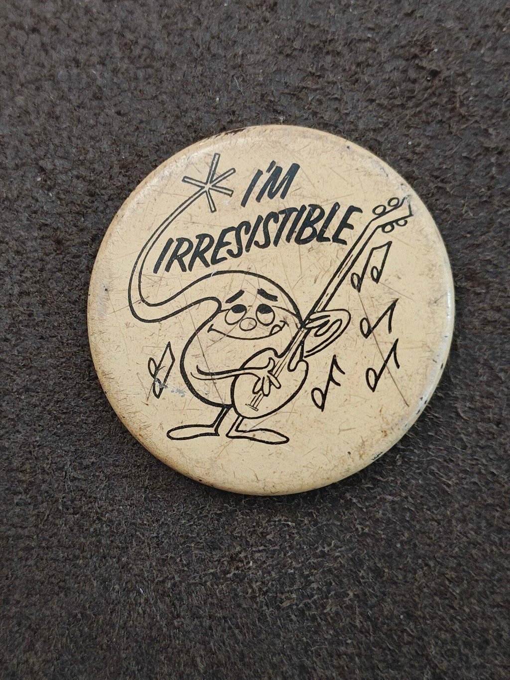 Round beige button pin with "I'm Irresistible" cartoon character and musical notes