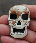 Skull-shaped enamel pin with brown-and-white face, black eyes, and white teeth