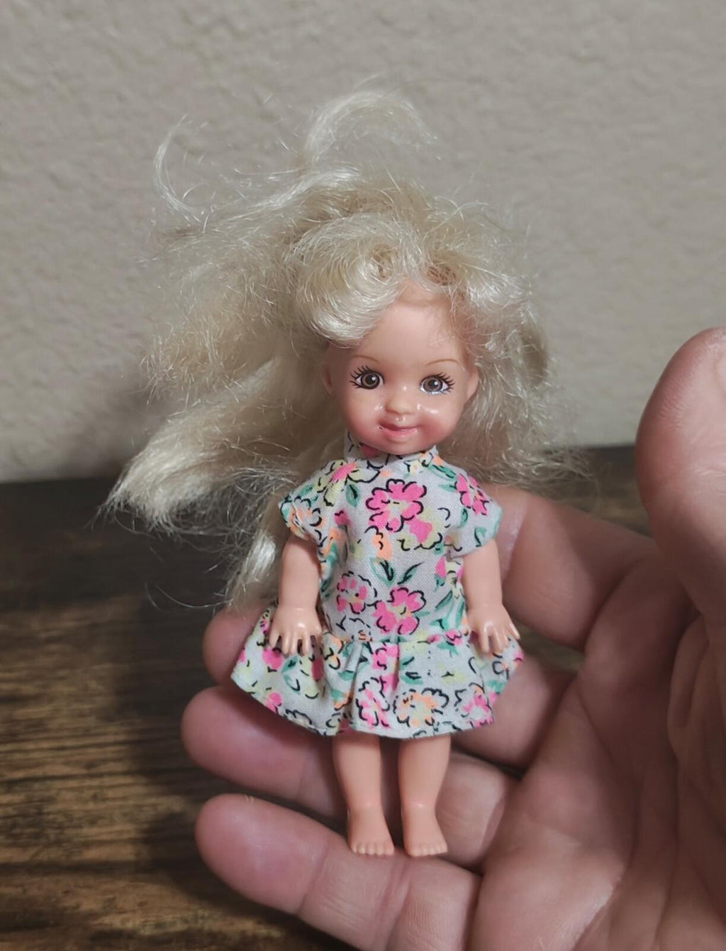 Mini girl doll with long curly blonde hair and floral pink-blue dress, held in hand.