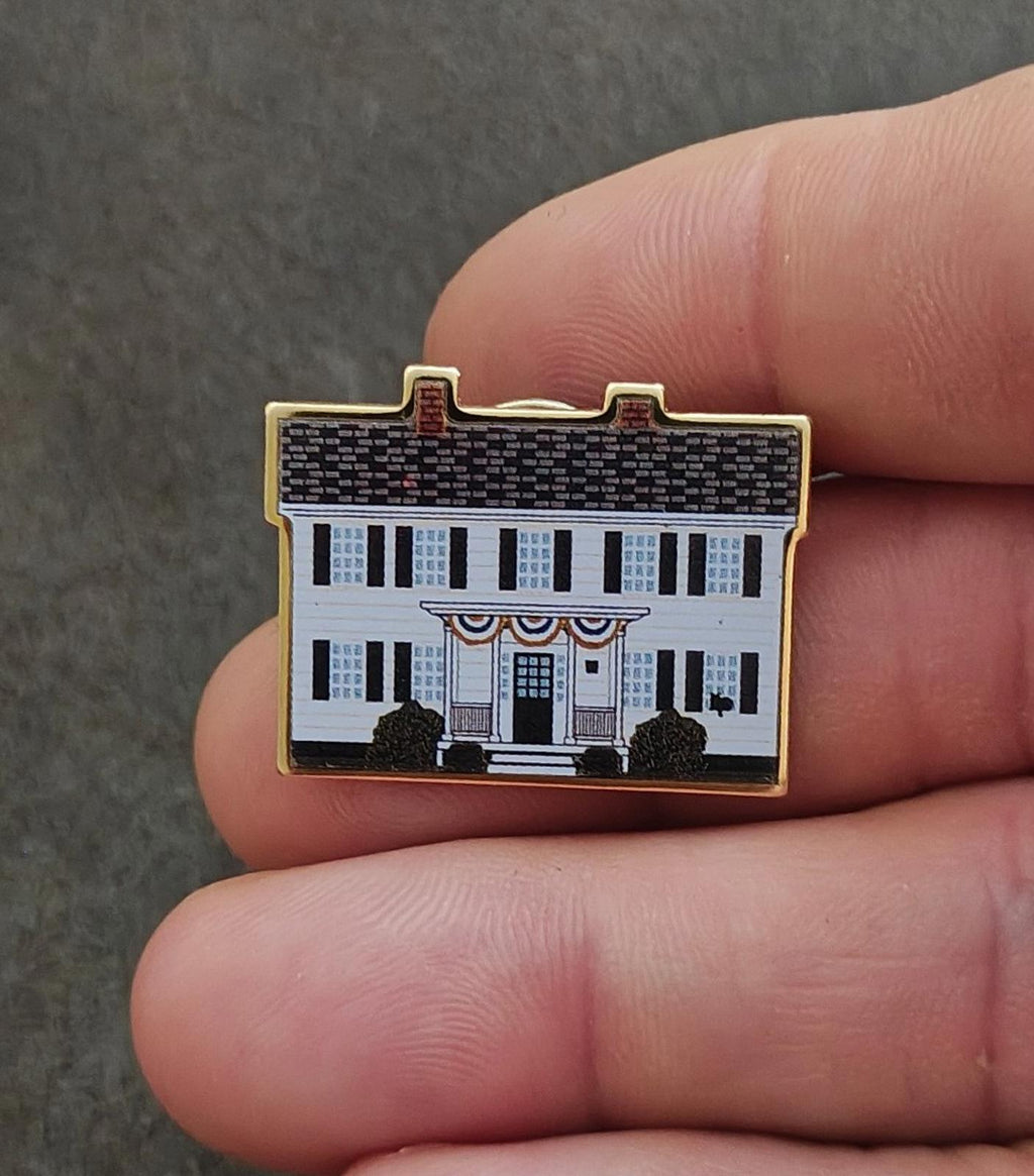 Gold-rimmed enamel pin held in fingers, featuring white building, blue windows, and trees