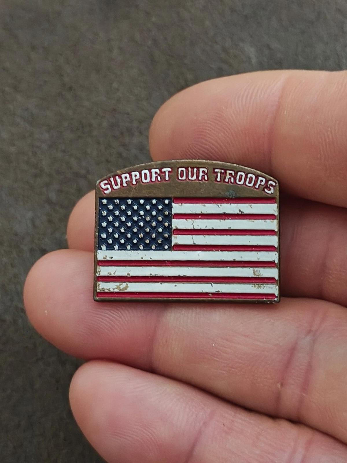 Gold-tone enamel pin held in hand, showing “Support Our Troops” over U.S. flag