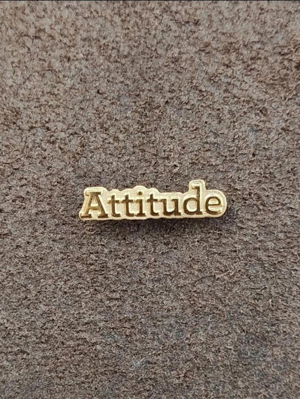 Gold tone lapel pin with raised "Attitude" letters on gray fabric