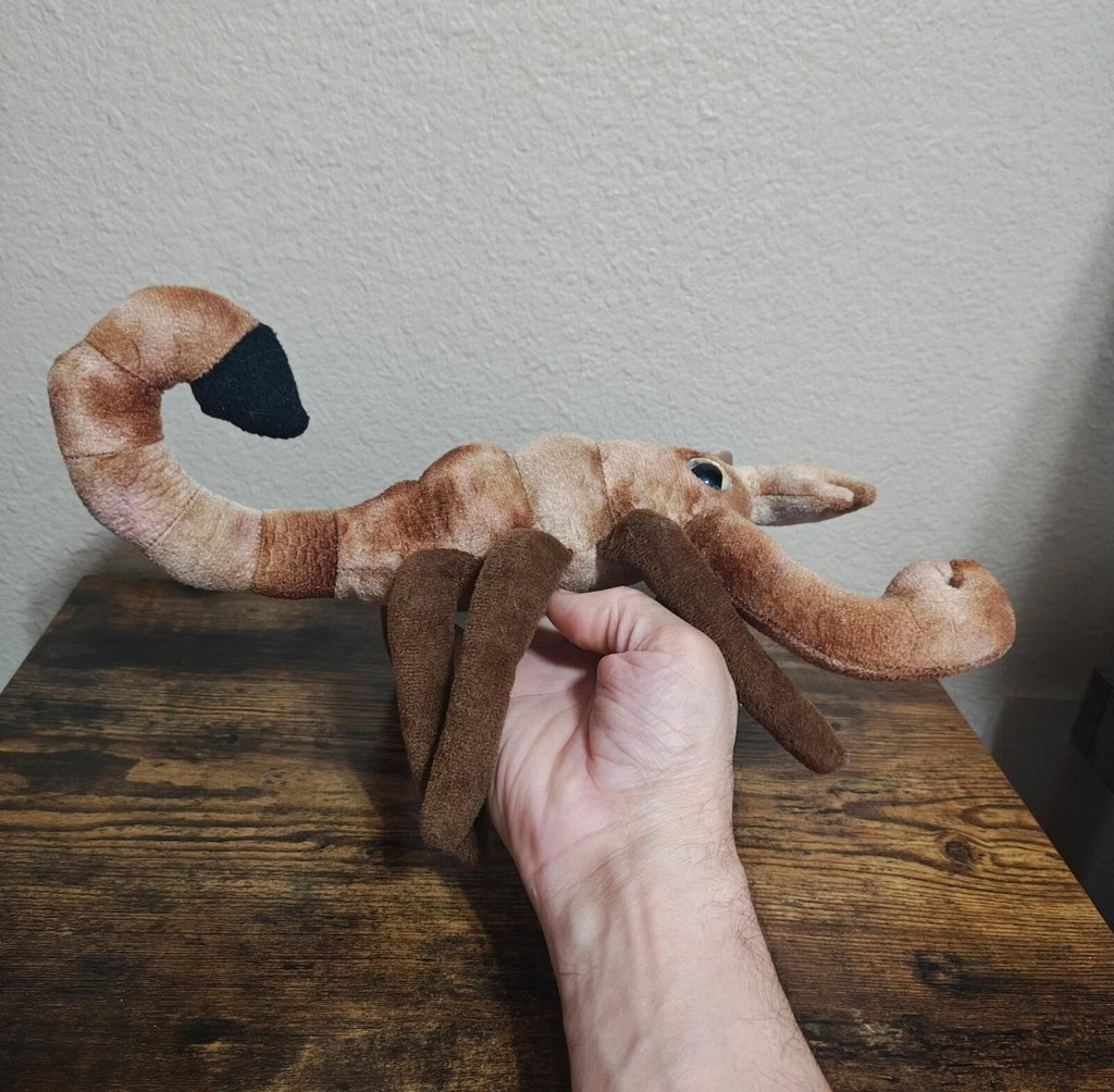 Brown plush scorpion stretched out on table, long tail and legs, held by hand