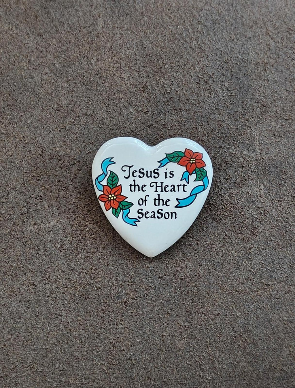 White ceramic heart pin showing “Jesus is the Heart of the Season” with blue ribbons.
