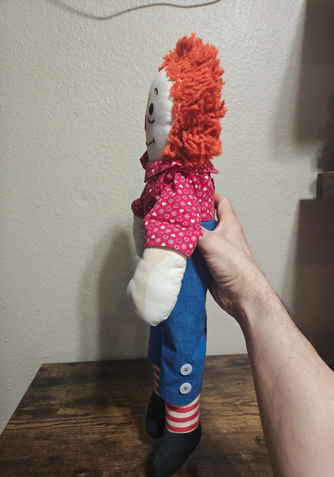 Handmade rag doll in side view wearing red heart-pattern shirt and blue overalls with striped socks.