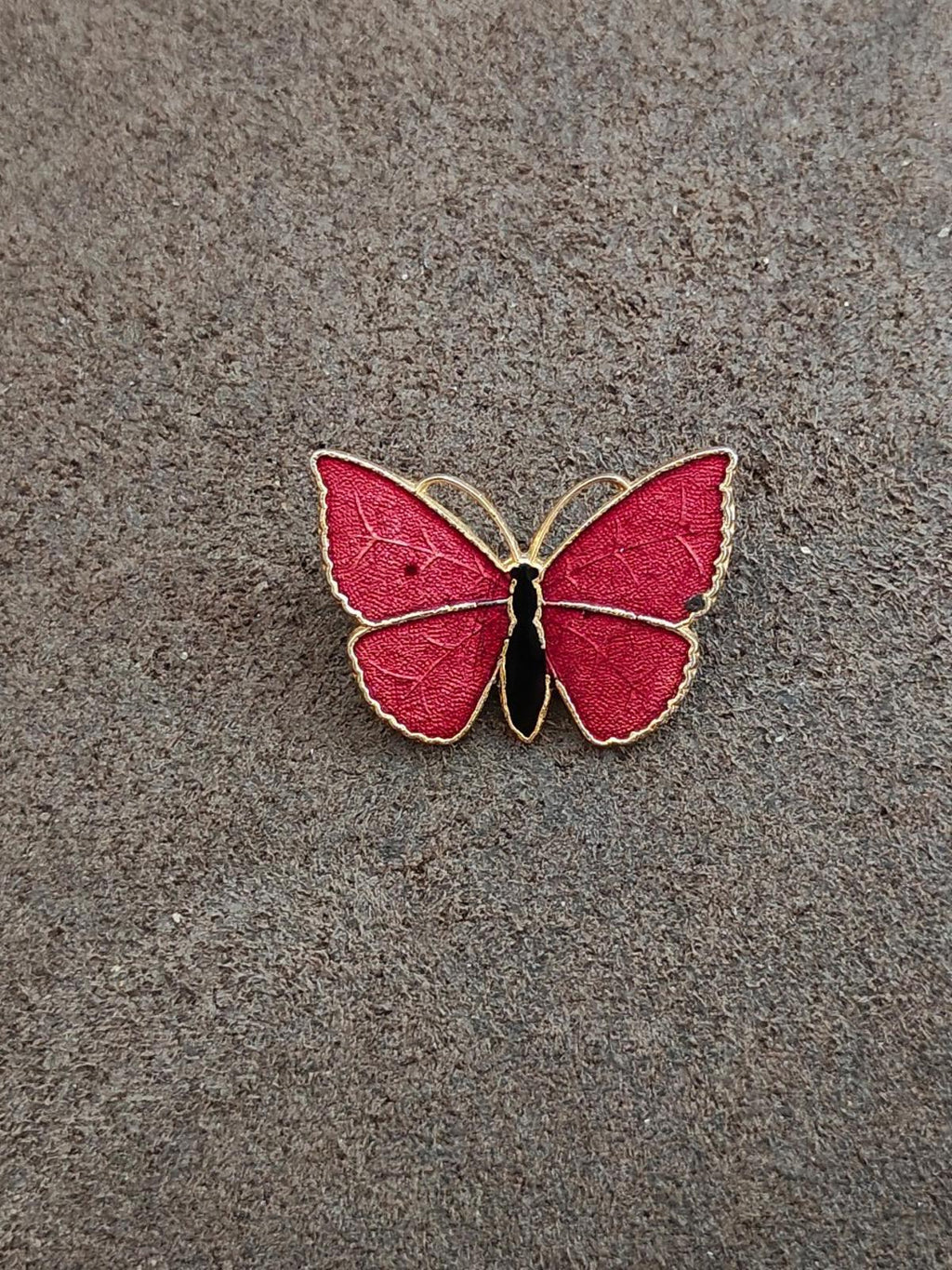 Red butterfly enamel brooch with gold-tone outline and black body, small on textured surface.