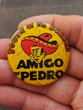 Yellow AMIGO OF PEDRO pinback button with sombrero mascot graphic, held in hand