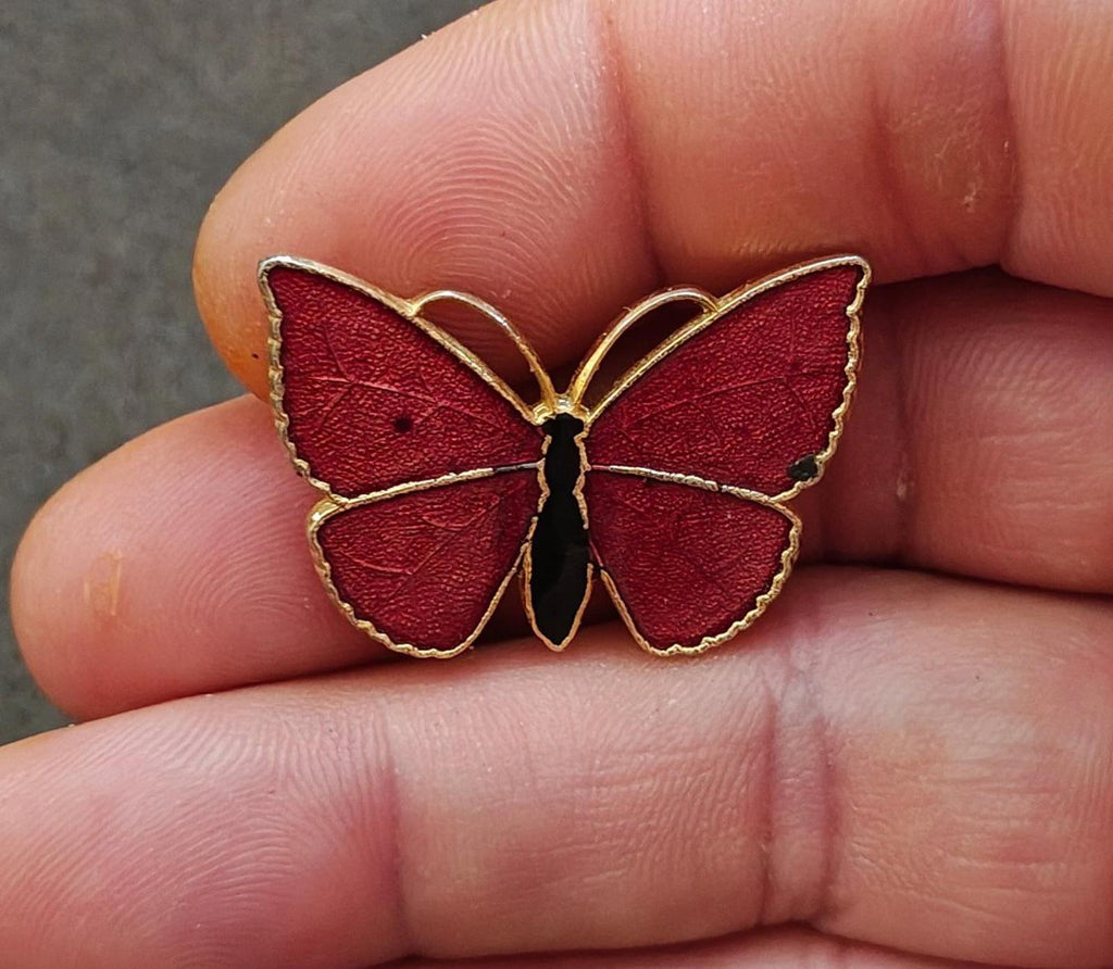 Red butterfly enamel brooch with gold-tone wing outline and black body, held in fingers.
