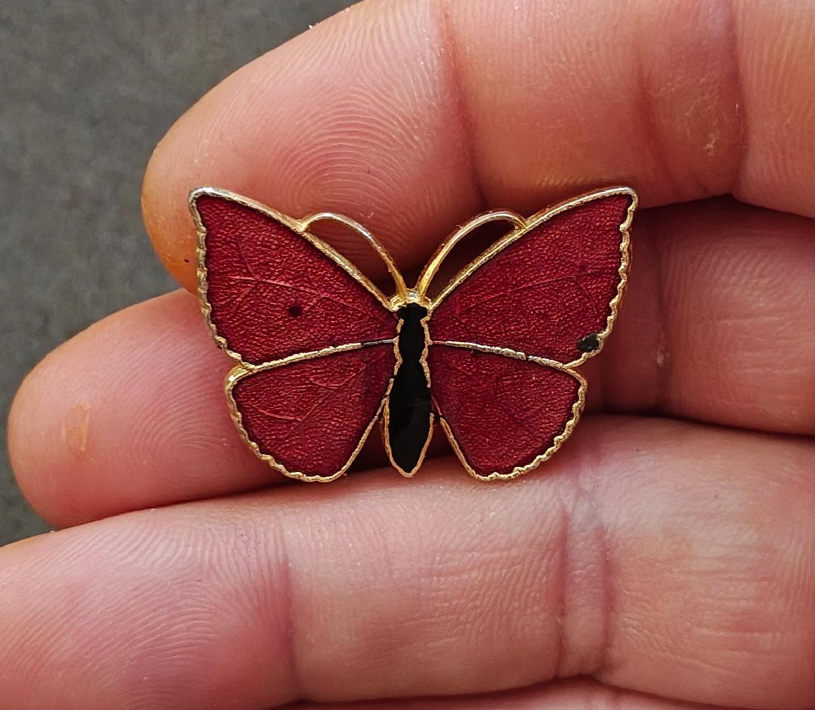Red butterfly enamel brooch with gold-tone wing outline and black body, held in fingers.