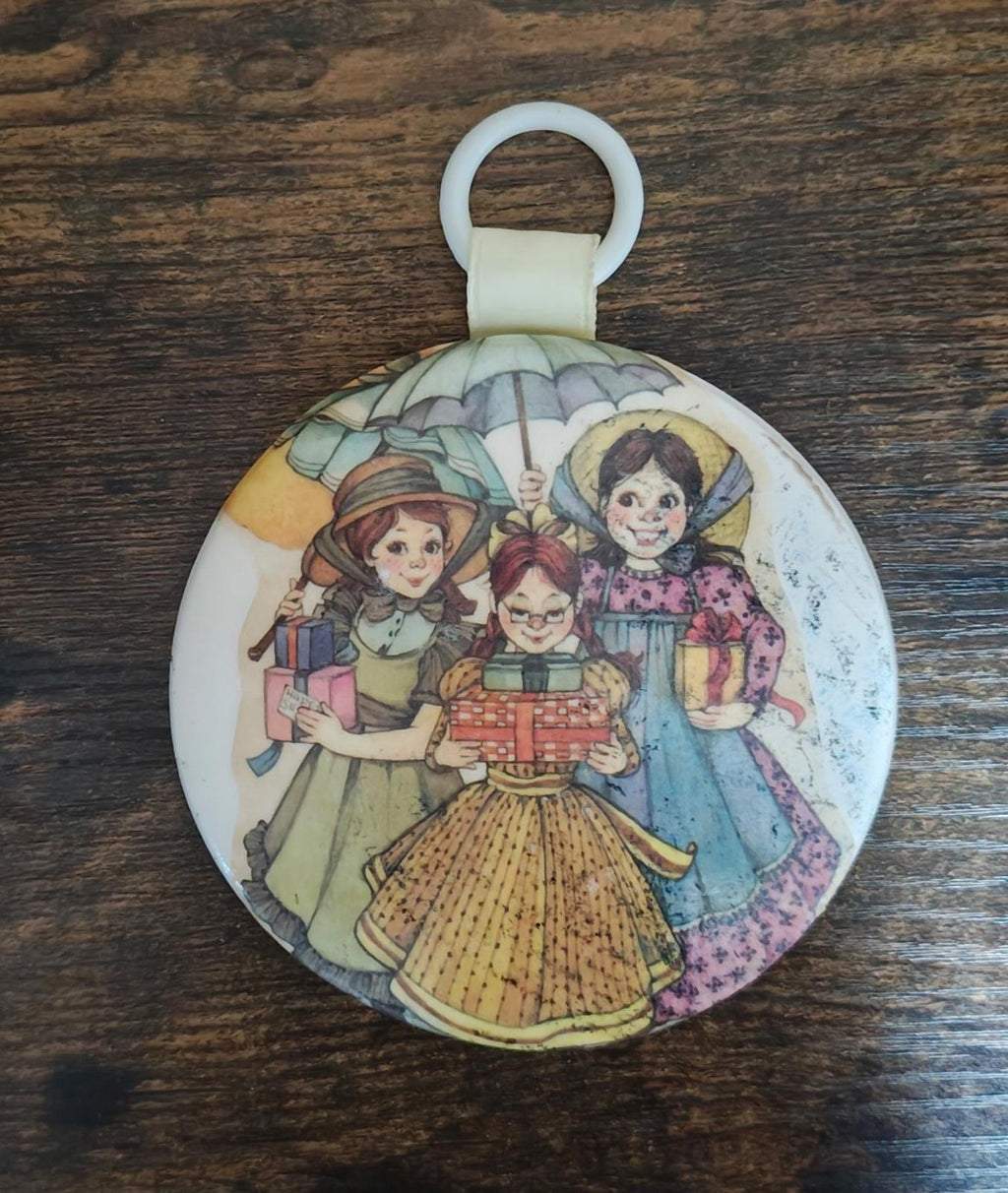 Round ornament featuring three illustrated girls and a small toy cart, cream loop