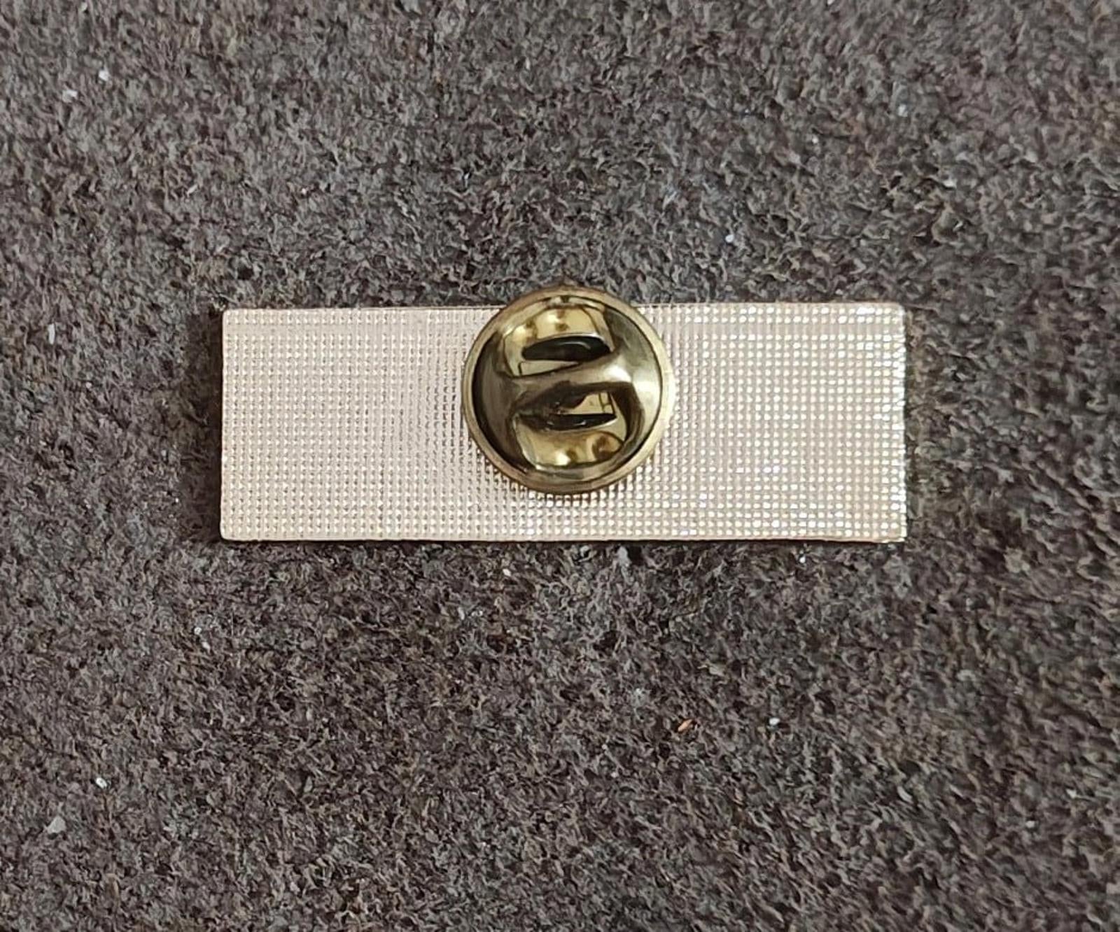 Gold-tone pin back with round clasp on a white backing rectangle.