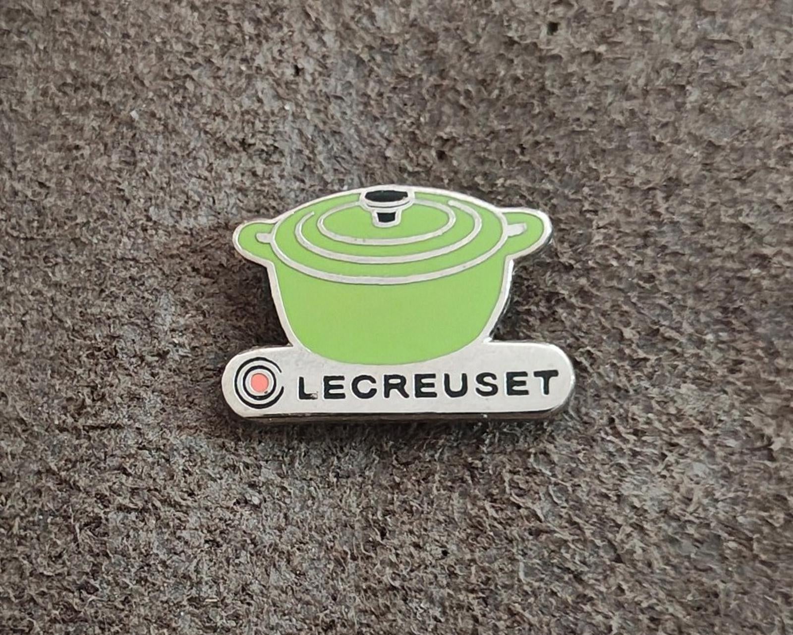 Le Creuset green mini cocotte enamel pin on gray fabric, straight-on view