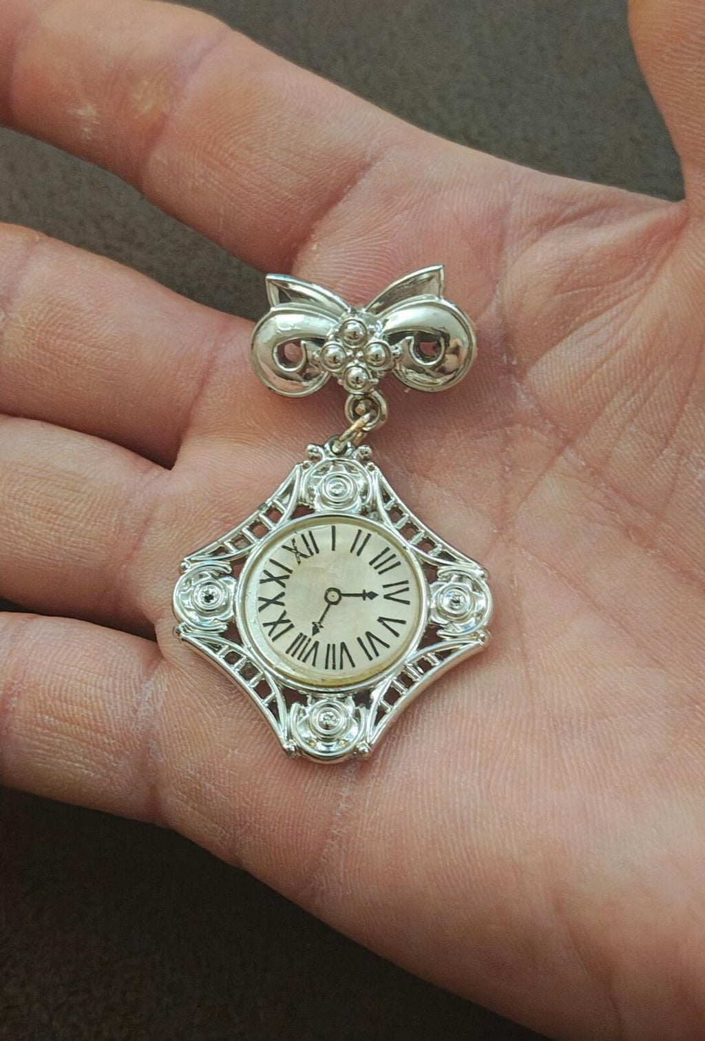 Silver-tone bow clock brooch on hand; decorative square clock face with roman numerals.