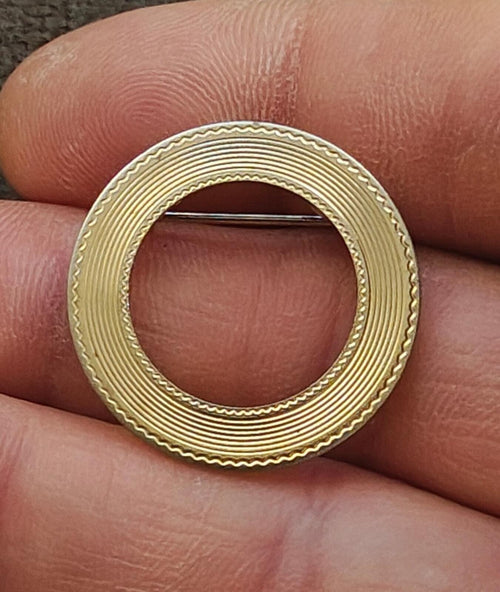 Gold textured circular brooch with central cutout and metal pin bar across the opening