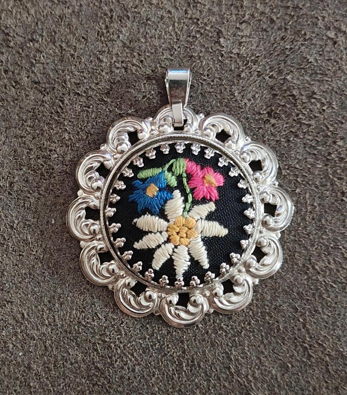Silver filigree round pendant with black fabric and stitched white and colored flower.