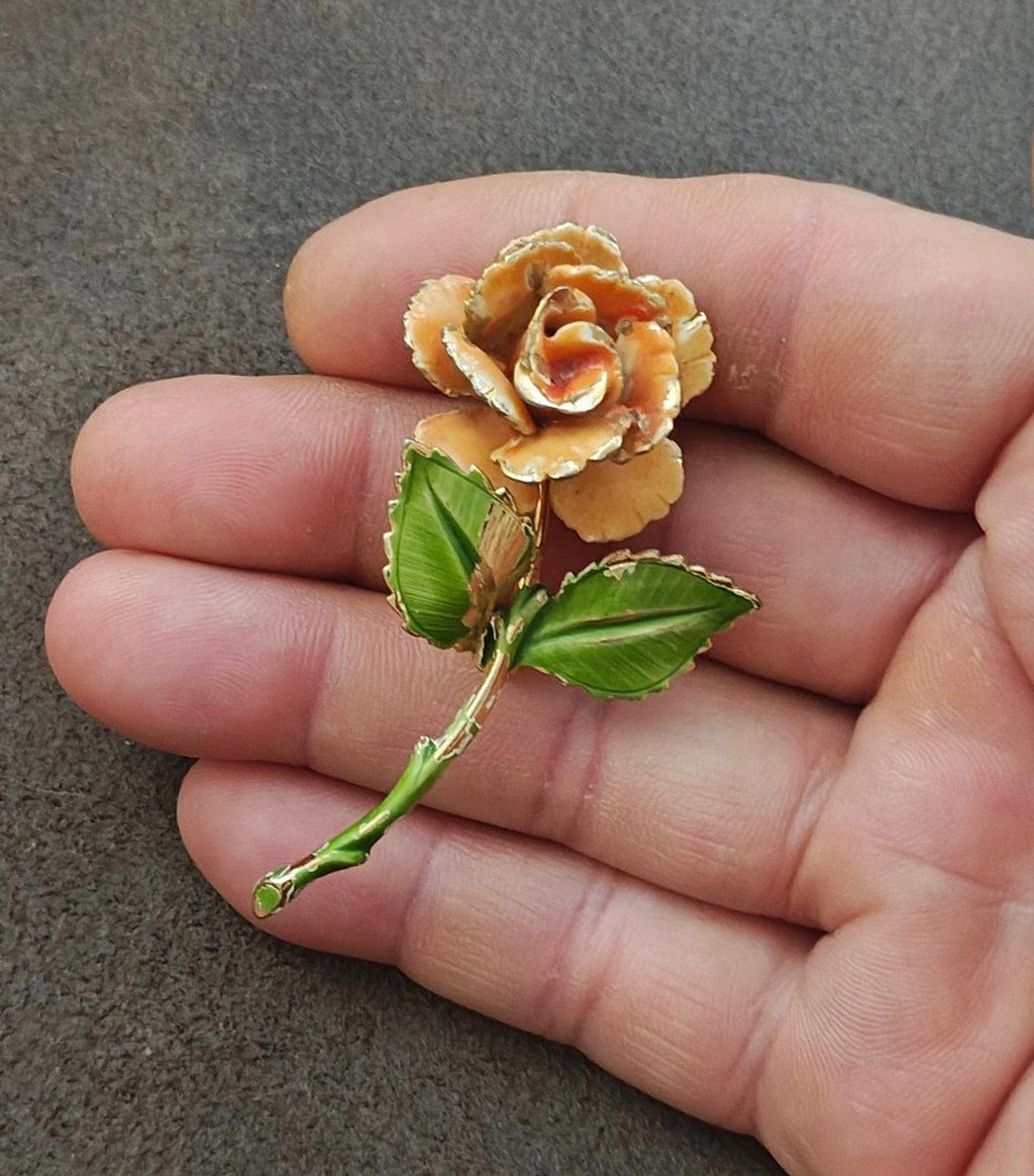 Gold-tone enamel rose pin with orange petals and green leaves, close-up on fingers
