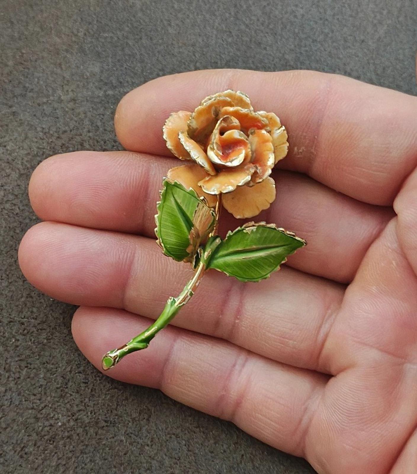 Gold-tone enamel rose pin with orange petals and green leaves, close-up on fingers