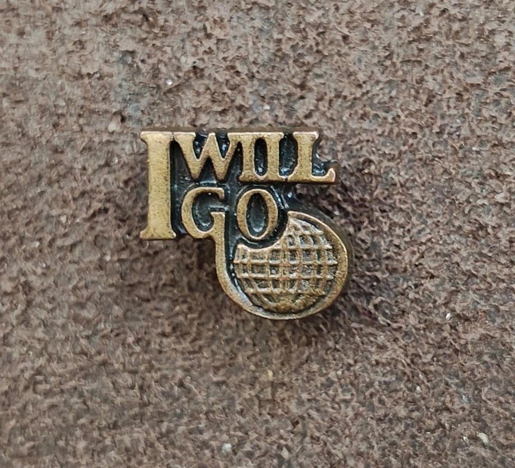 Gold tone "I WILL GO" lapel pin with globe-style emblem, front view on textured surface