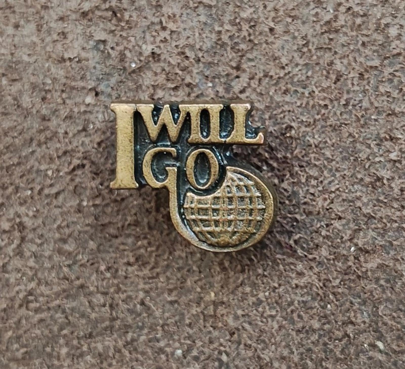 Gold tone "I WILL GO" lapel pin with globe-style emblem, front view on textured surface