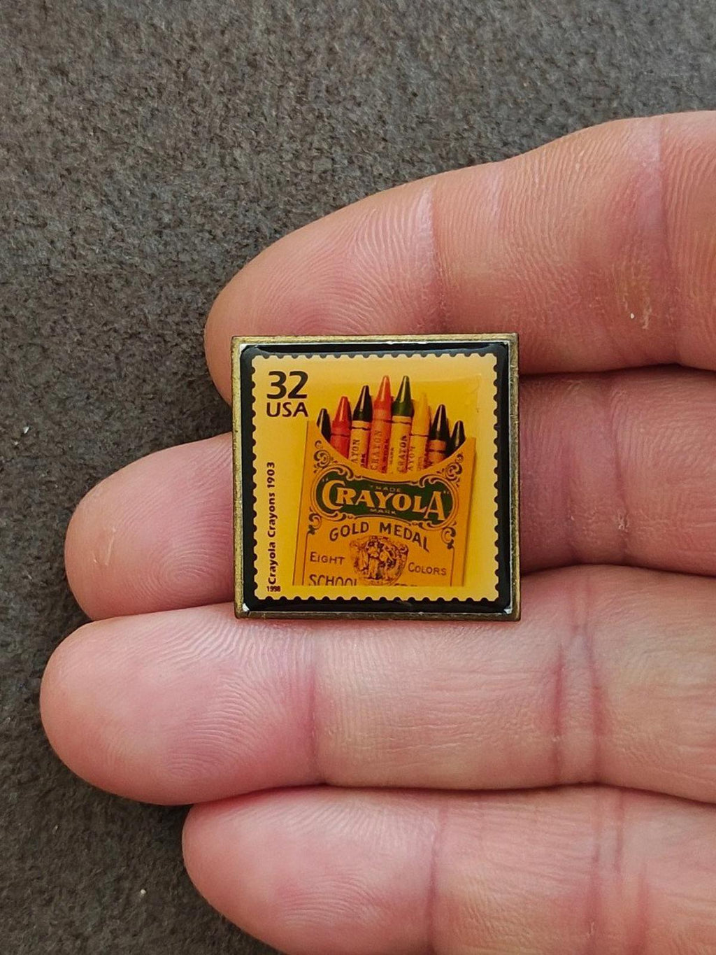 Square gold-tone pin showing “32 USA” and Crayola Gold Medal text with eight crayons