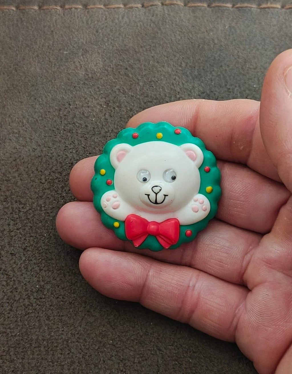 Green teddy bear wreath pin in hand, white bear face with red bow and googly eyes