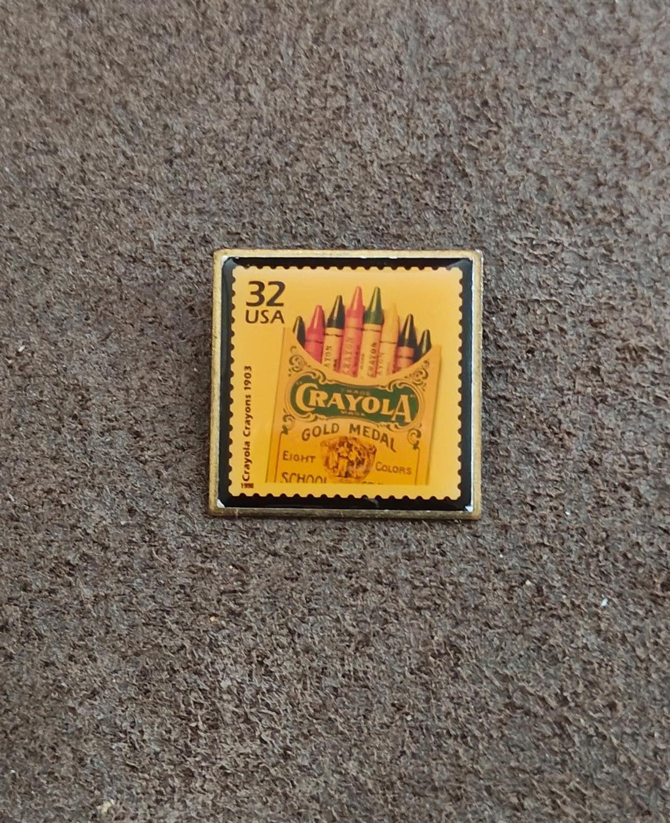 Square gold-tone pin with yellow “32 USA” stamp design and Crayola Gold Medal crayons