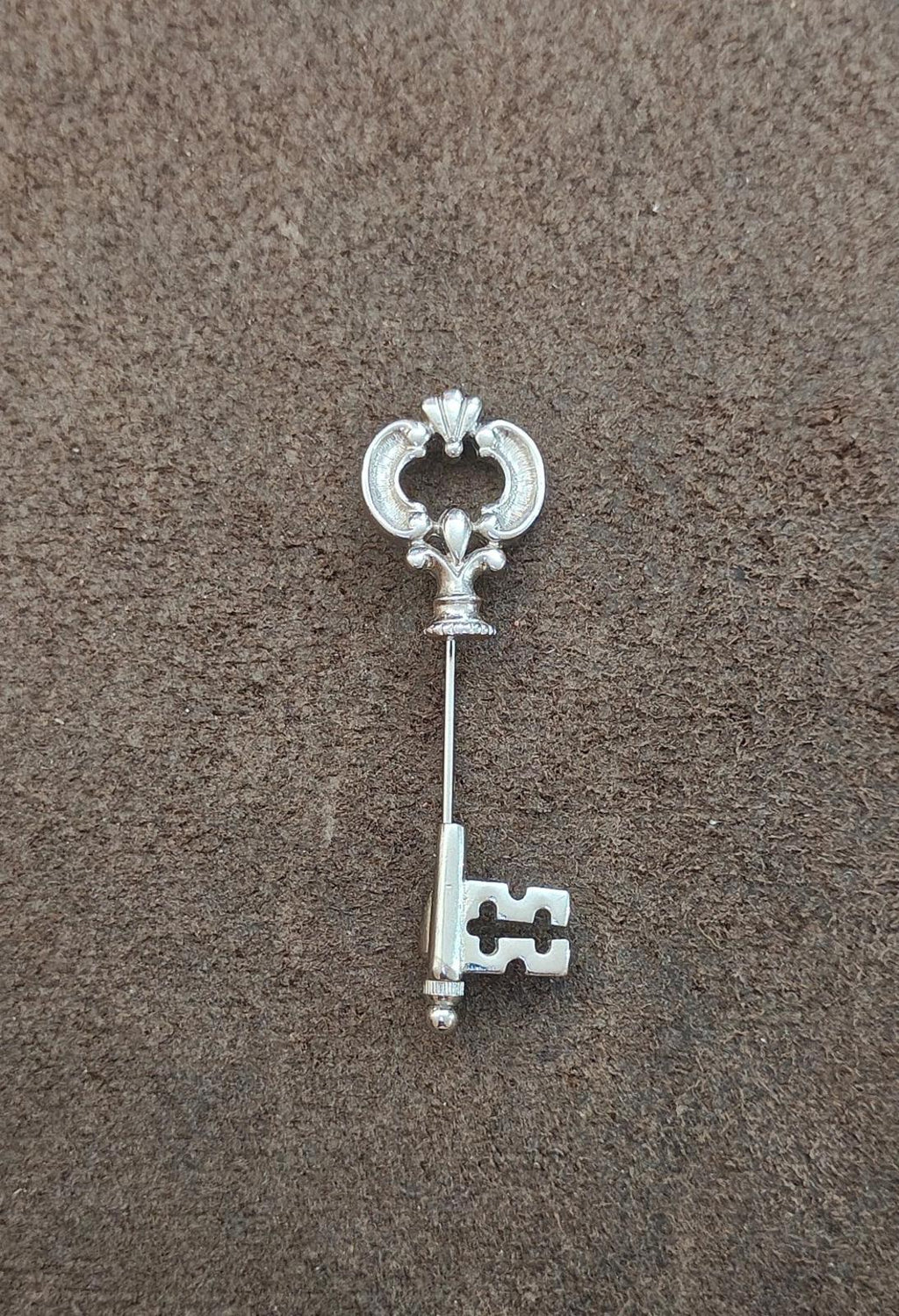 Silver skeleton key stick pin with fleur-de-lis head, shown from above on dark surface.