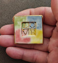 Square polymer clay brooch with gold-tone border and 'LOVE' letters on pastel background