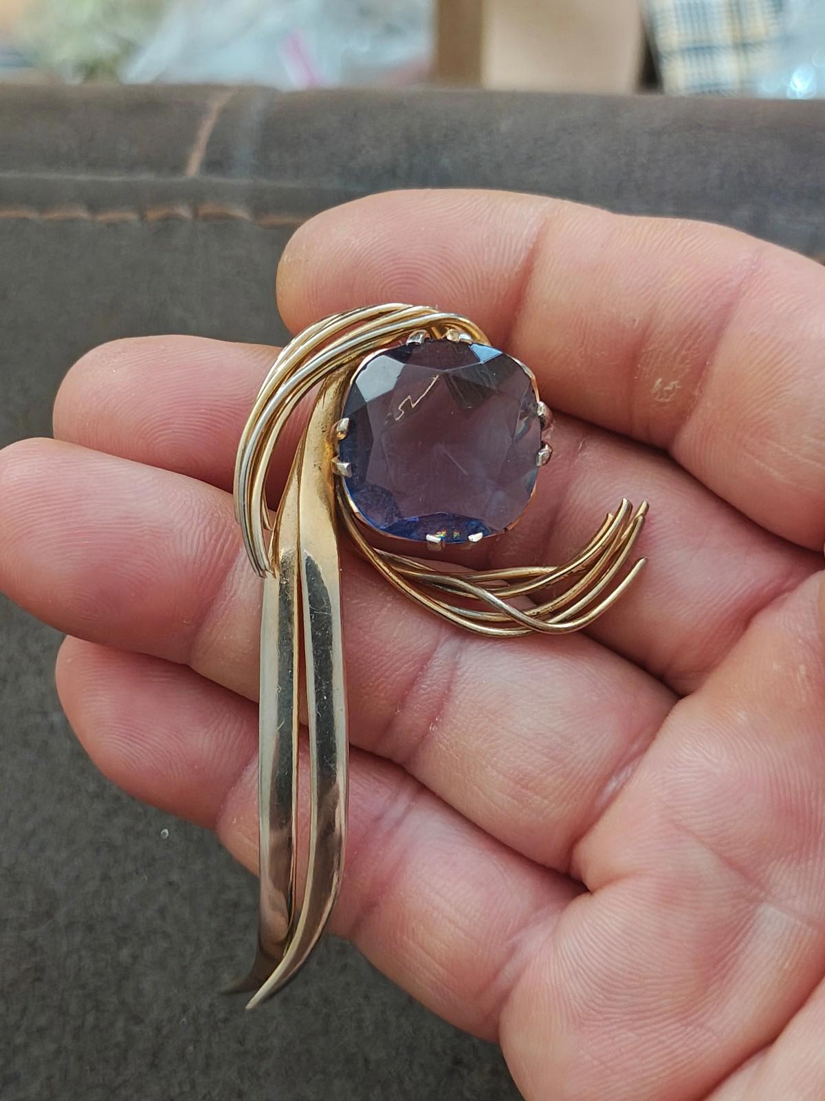 Gold-tone abstract brooch pin with blue gemstone in round setting, shown in hand