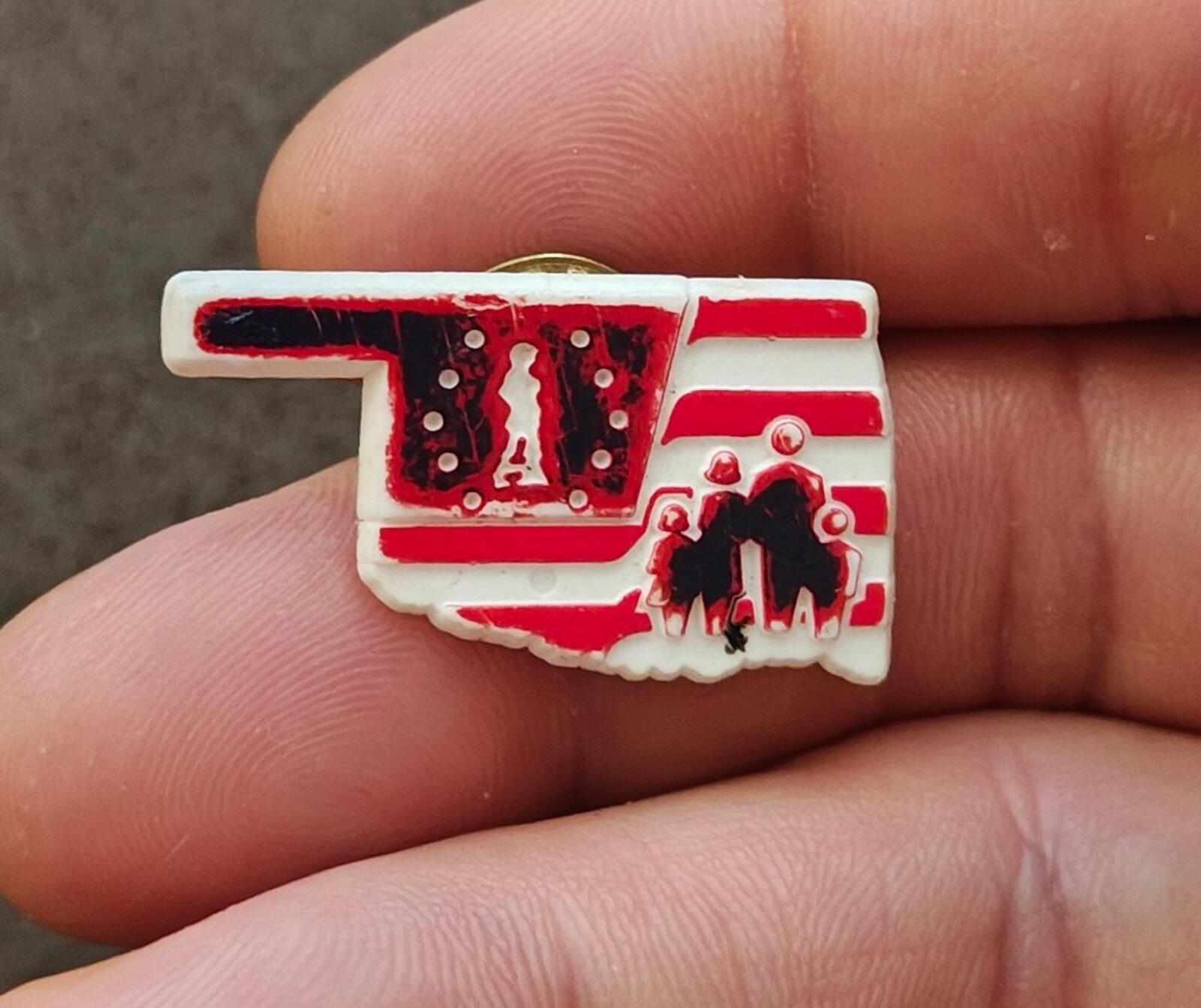 Red and white Oklahoma map plastic lapel pin, front view close-up in hand
