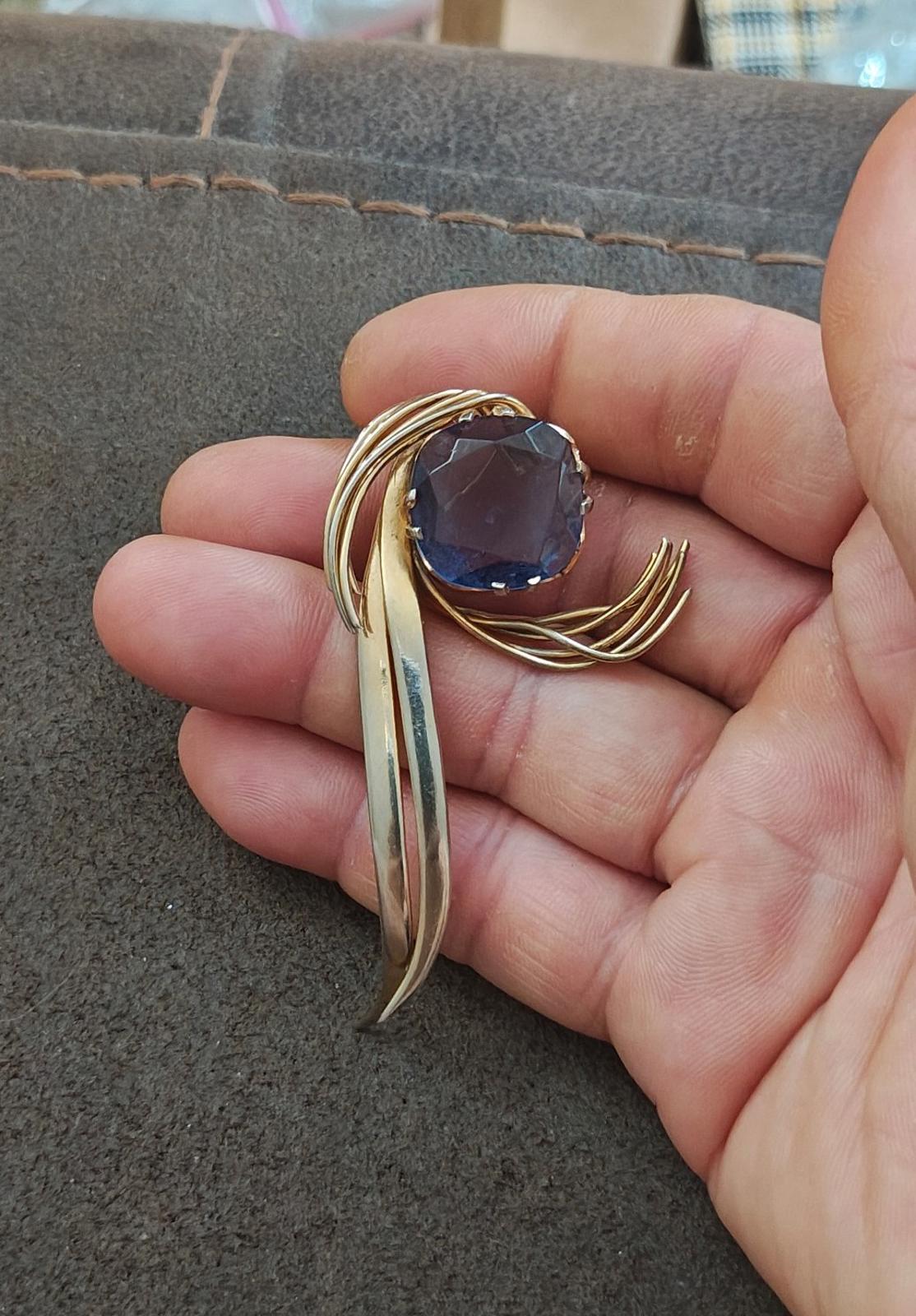 Gold-tone abstract brooch pin with blue gemstone, swirl edges, held on palm