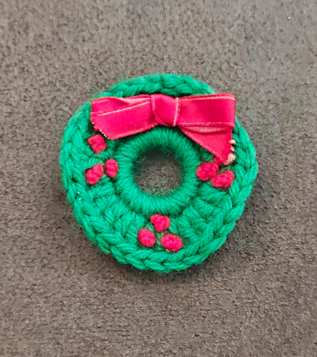 Green knitted wreath brooch with pink ribbon bow and red berry details, centered ring